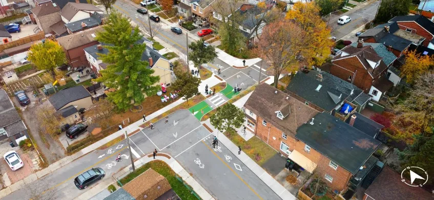 Street-level view of a rebuilt intersection with new bollards, a raised crosswalk, and a Do Not Enter Except Bicycles sign, houses and a convenience store in the background
