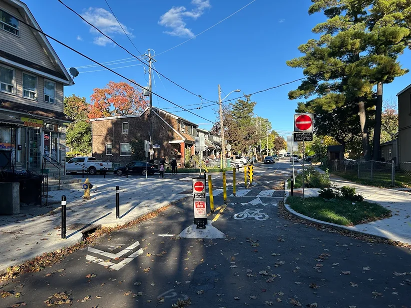 Street-level view of a rebuilt intersection with new bollards, a raised crosswalk, and a Do Not Enter Except Bicycles sign, houses and a convenience store in the background