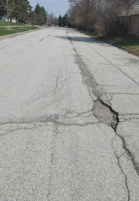 Ground-level view of a cracked, deteriorated asphalt road surface with visible patch repairs and longitudinal cracking
