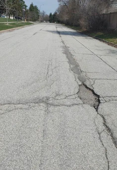 Ground-level view of a cracked, deteriorated asphalt road surface with visible patch repairs and longitudinal cracking