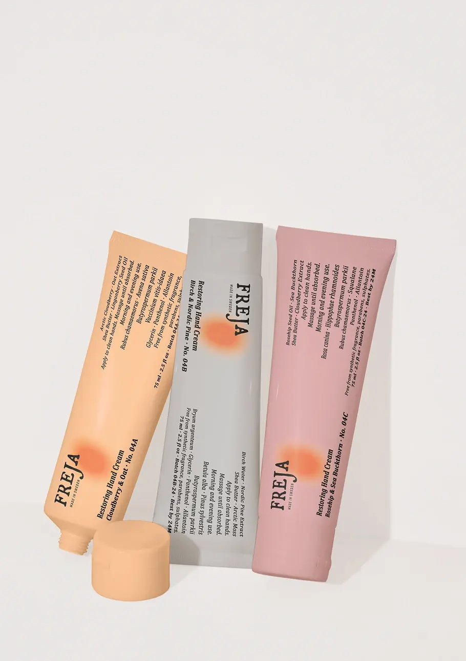 Three Freja product tubes in orange, gray, and pink lined up against a white background