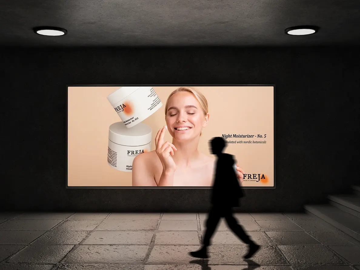 A person walking past a lit Freja ad panel for the night moisturizer in a dark hallway